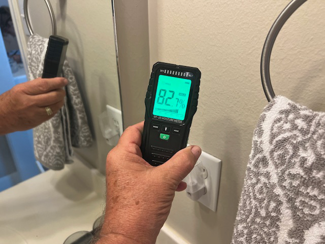 Person holding a black device displaying 82°F near a bathroom wall, with a mirror and towel rack visible.