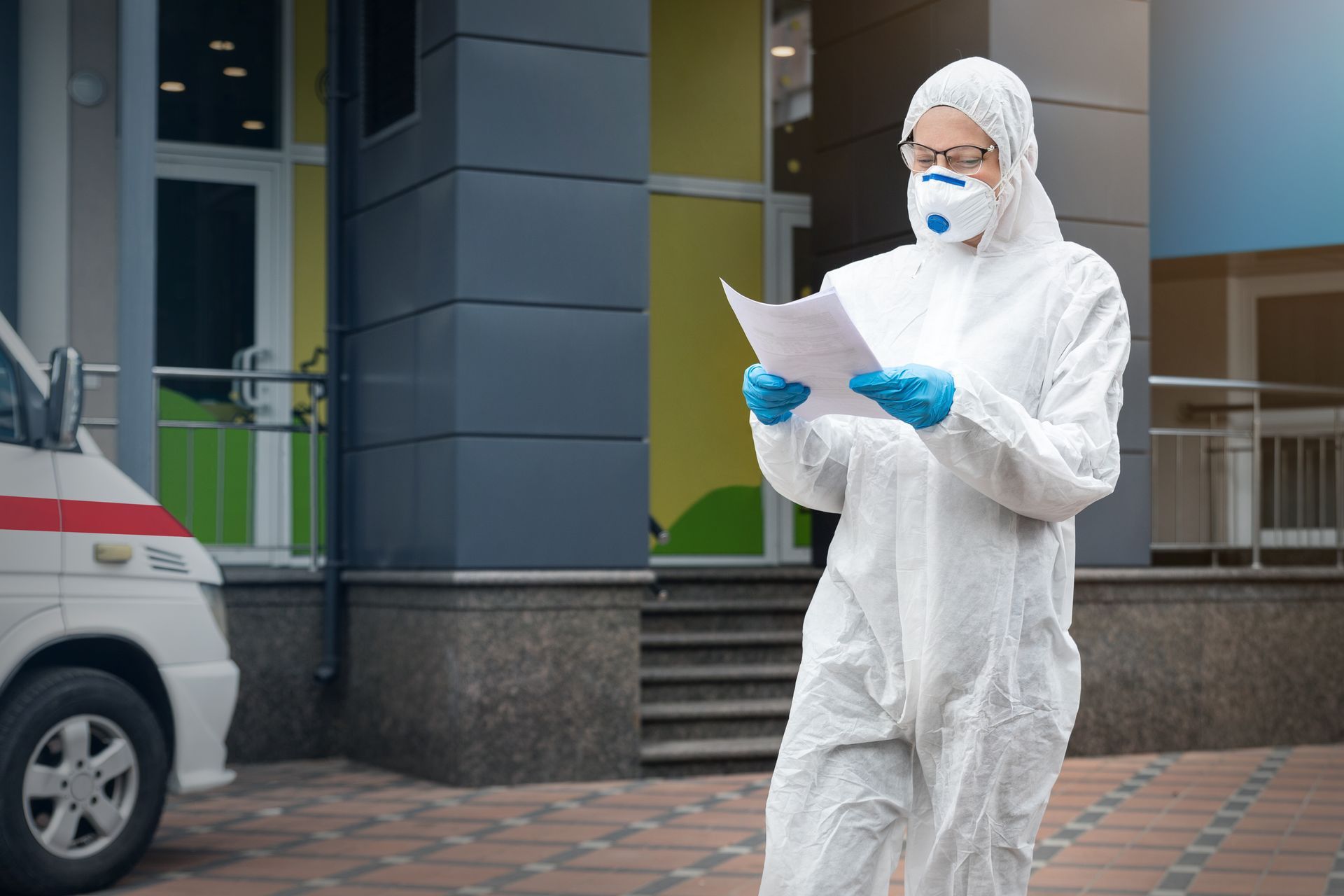 Person in white hazmat suit, mask, and gloves reads papers near ambulance and building entrance.