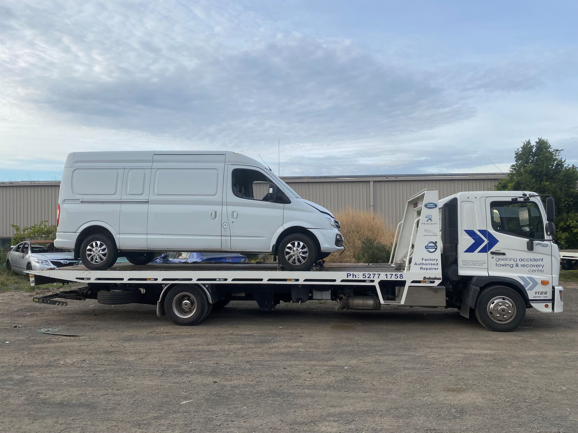 White van on a flatbed tow truck outdoors under a cloudy sky.