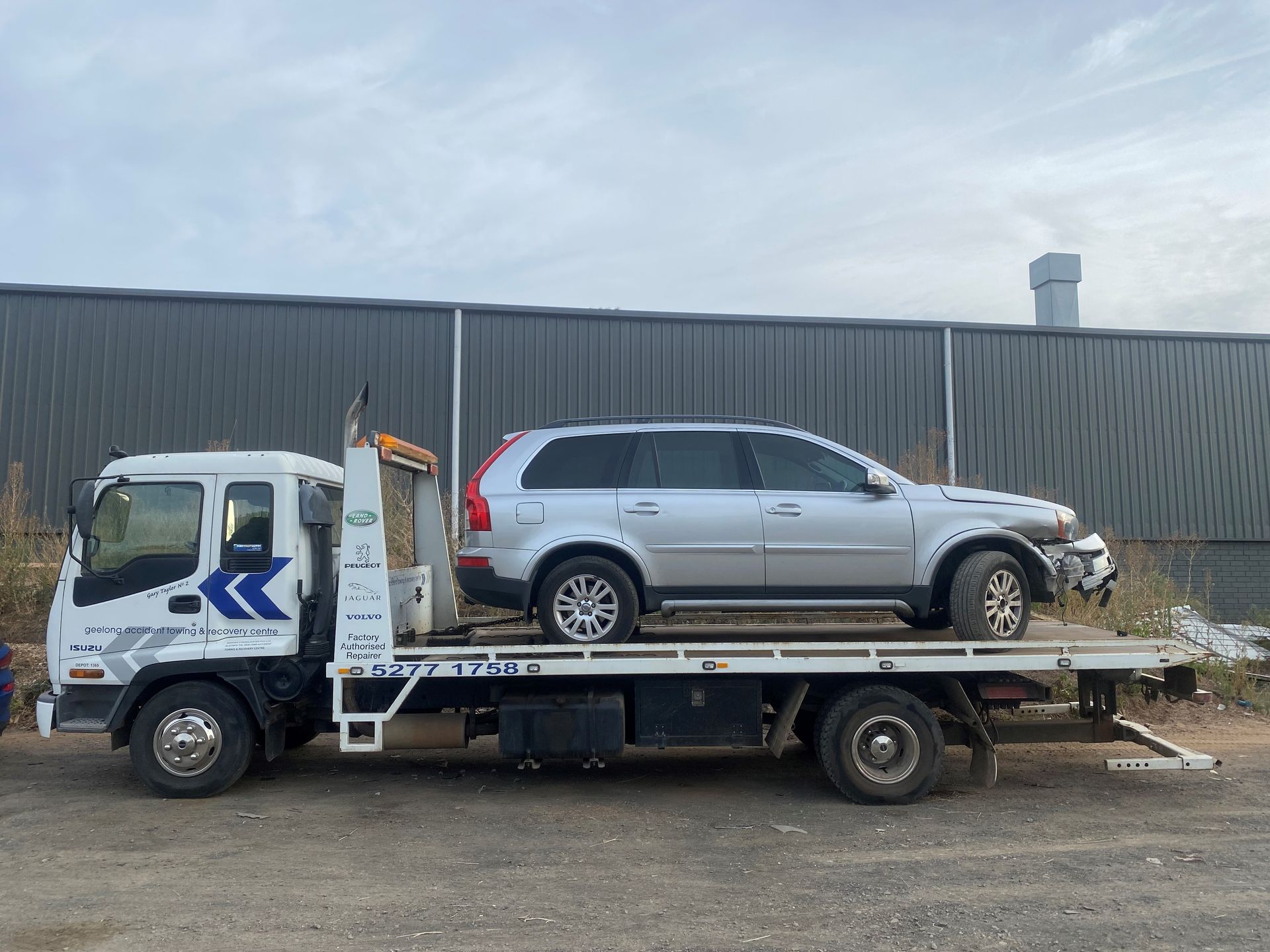 Tow truck carrying a silver Volvo SUV with front-end damage; parked in front of a gray industrial building.