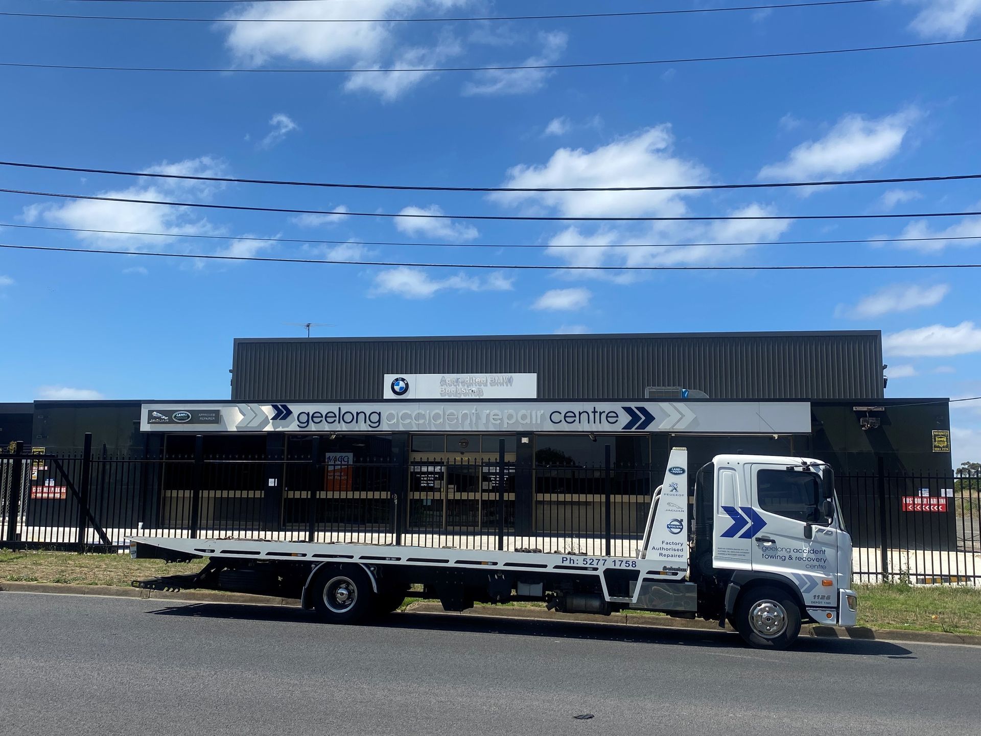 Flatbed tow truck parked in front of a black building with a sign on a sunny day.