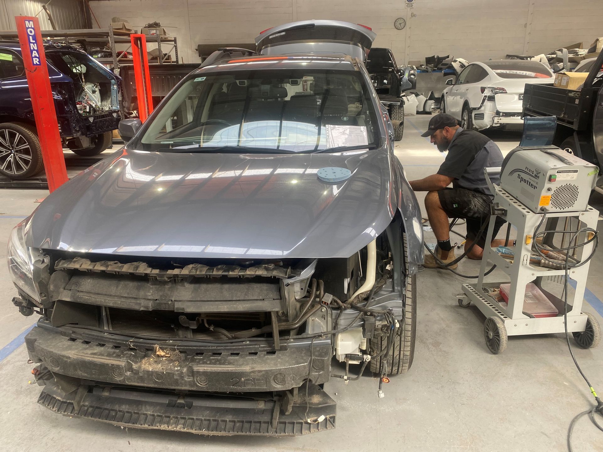 Car in a repair shop with front damage; a worker uses a machine to work on it.