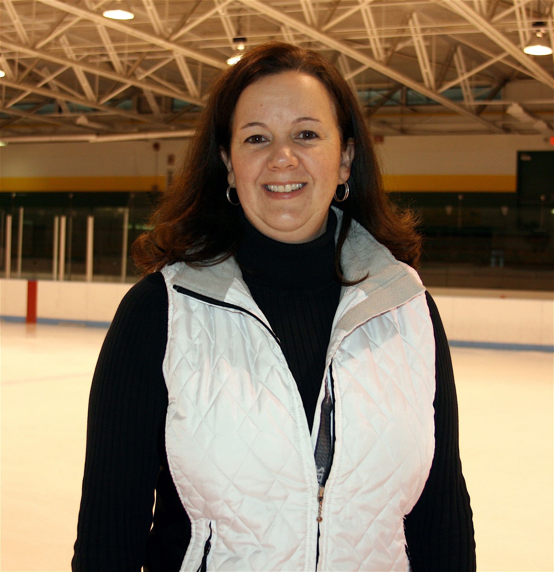 Woman in a white vest standing on an indoor ice rink, smiling at the camera.