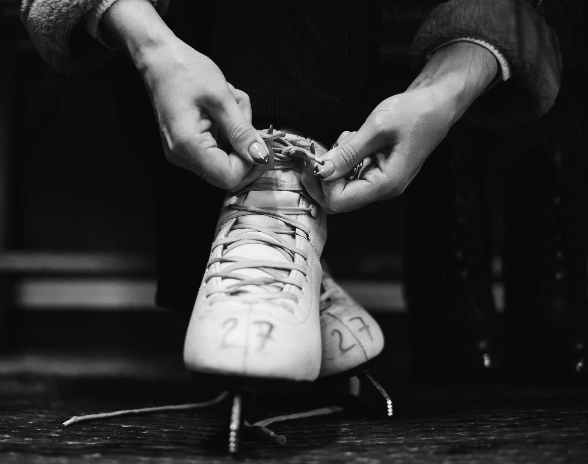 Hands tying laces on white ice skates with the numbers 27 and 23 on the blades.