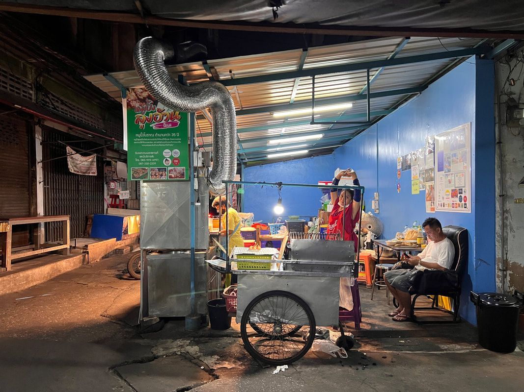 A street food vendor cooking, with a man seated nearby. Shop is under a blue awning at night.