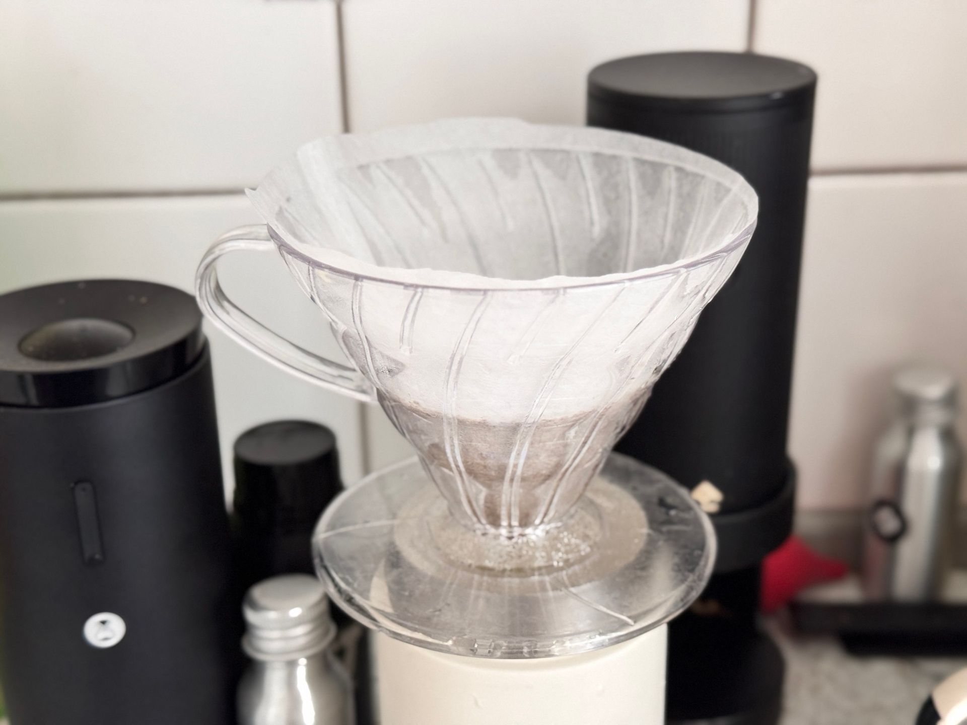 Pour-over coffee setup with filter in clear glass dripper, on a white scale, black grinder in background.