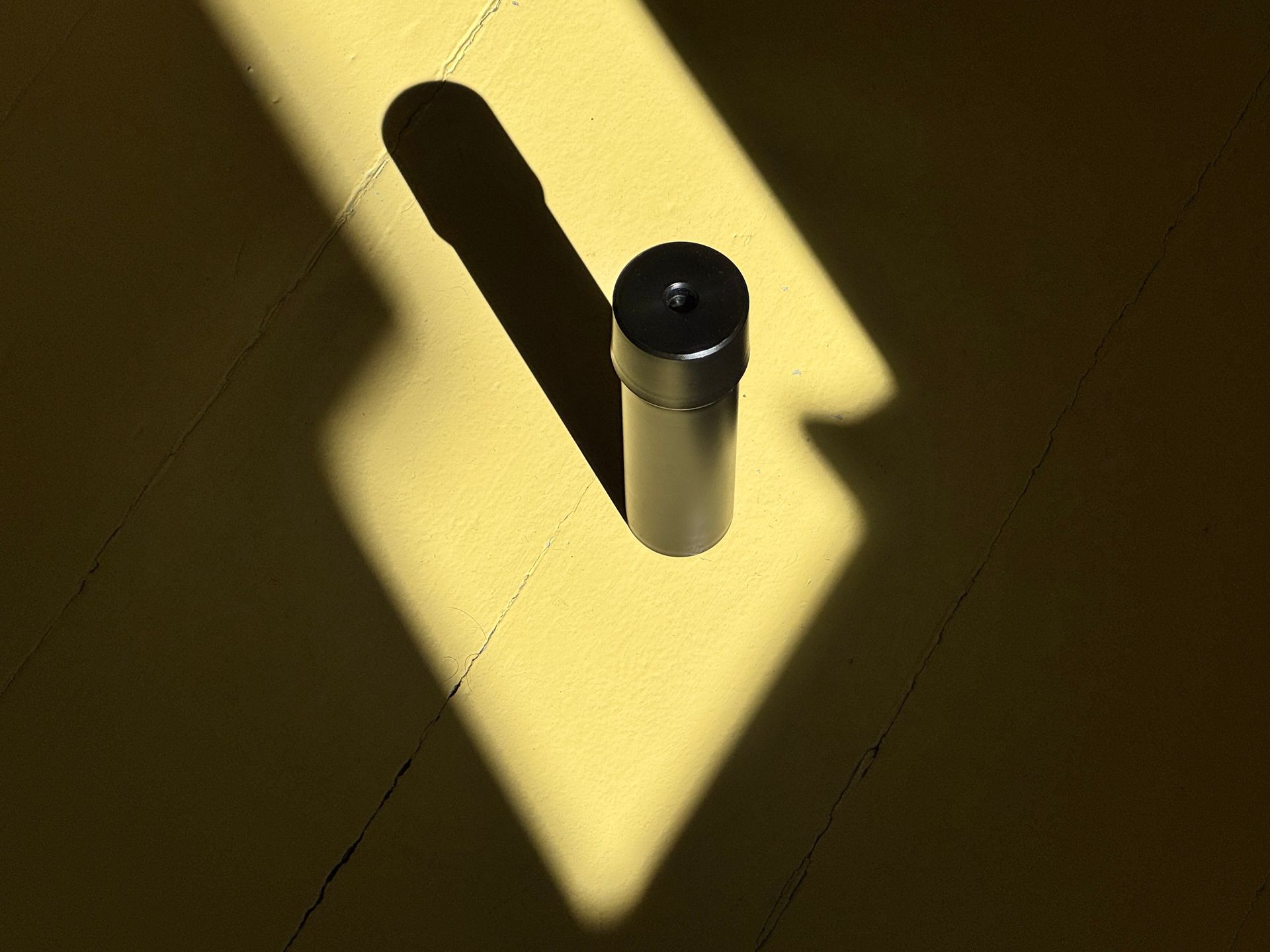 Black cylindrical object casting a long shadow on a sunlit yellow floor