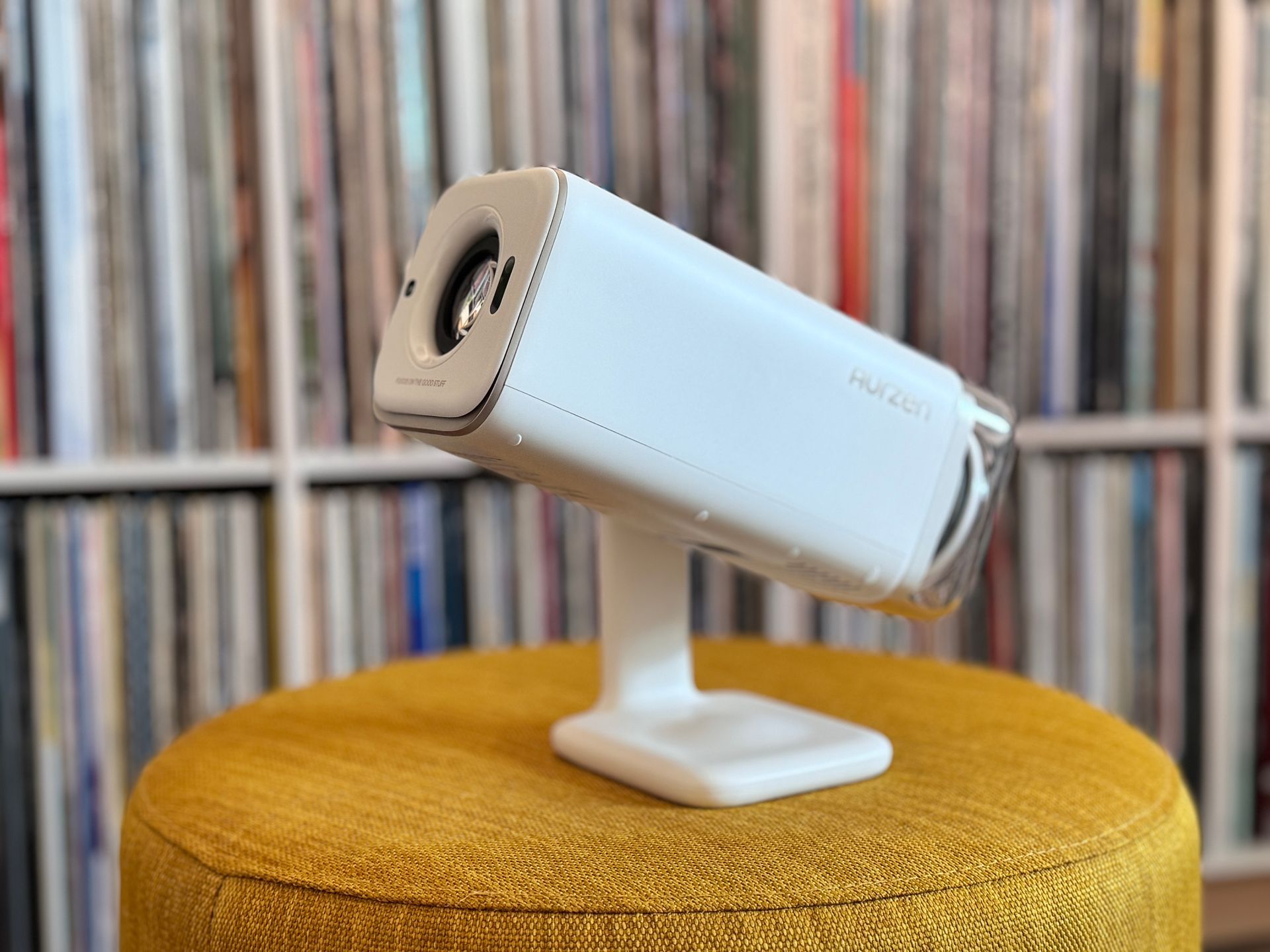 White security camera on a stand atop a mustard-yellow stool, with a blurred record shelf background