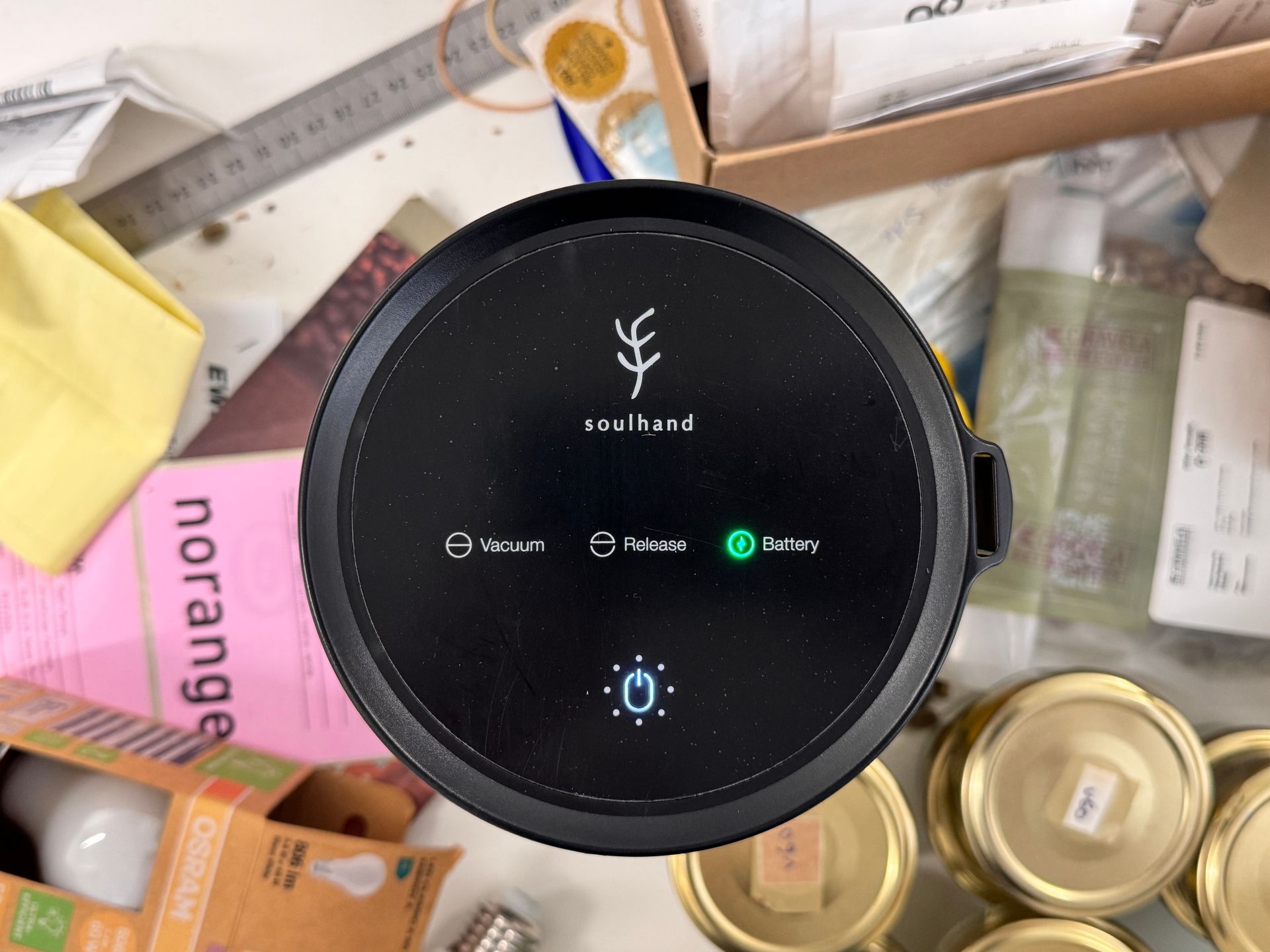 Circular black smart device on a cluttered desk, showing a central logo and small app icons on its screen.
