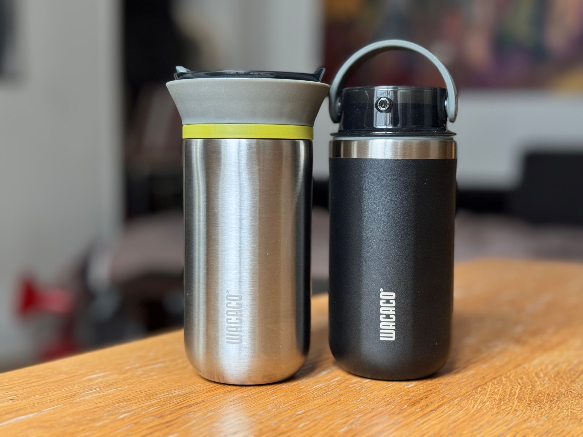 Two insulated travel mugs, one silver with a yellow lid and one black with a handle, on a wooden table.