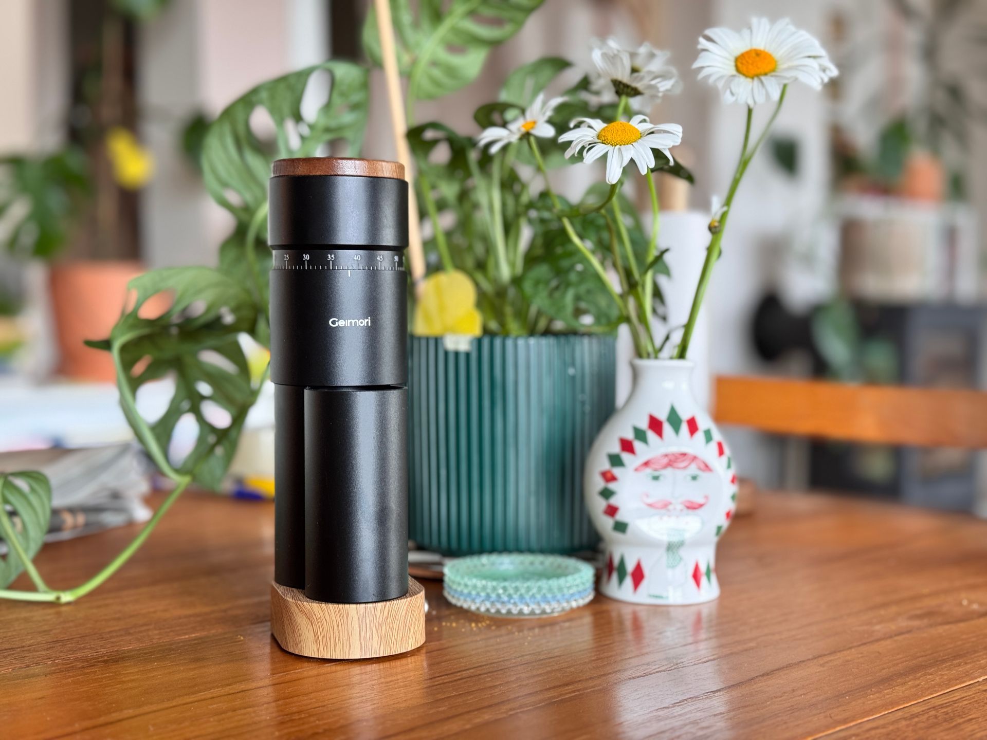 Black portable coffee grinder on wooden table with flowers in vase and small white figurine