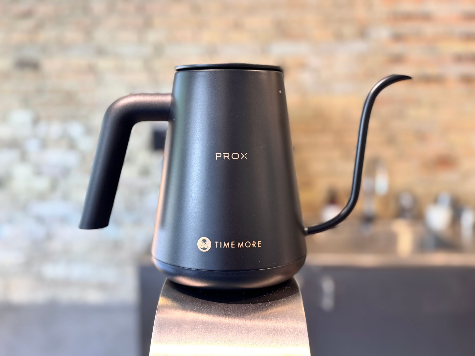 Black gooseneck electric kettle on a wooden base against a blurred brick kitchen wall