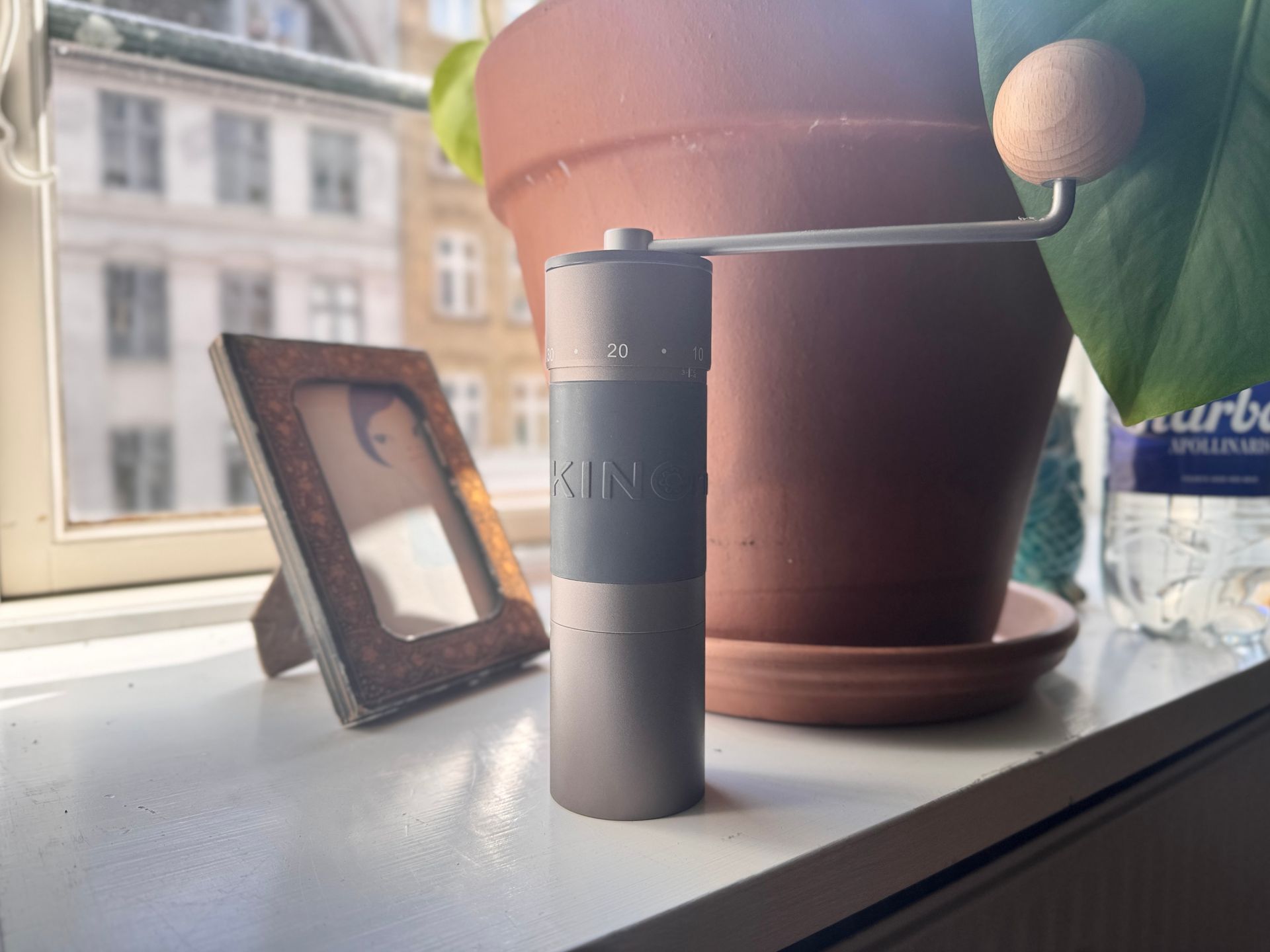 Manual coffee grinder on a windowsill, with a potted plant and a framed photo in the background.