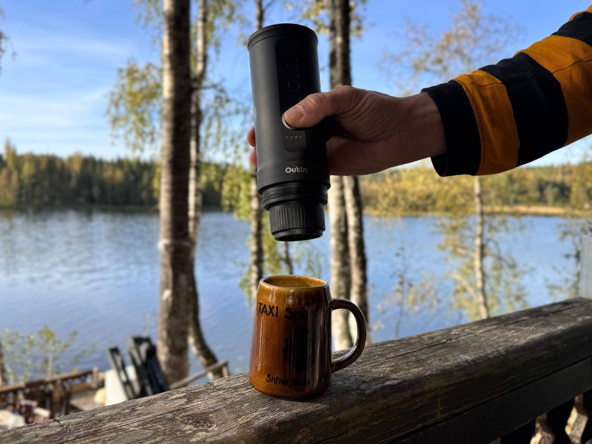 Person pouring from a black travel coffee maker into a brown mug, lakeside setting.