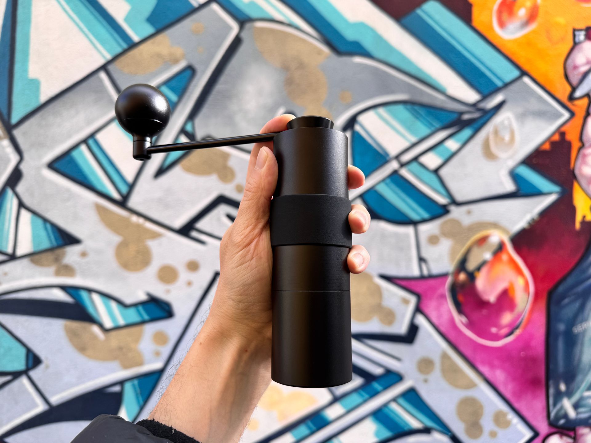 Person holding a black manual coffee grinder against a colorful graffiti background.