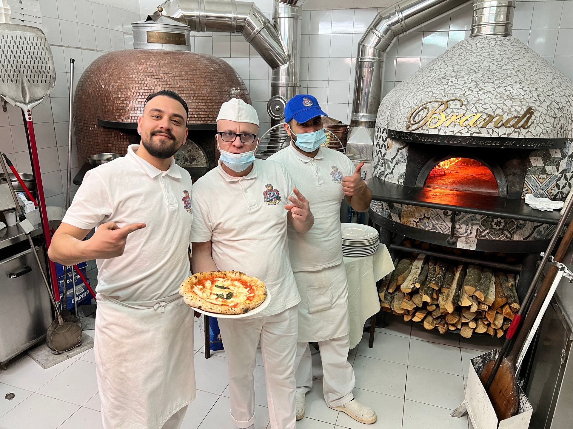 Three pizza makers by wood-fired ovens, one holding a pizza. Restaurant kitchen setting.