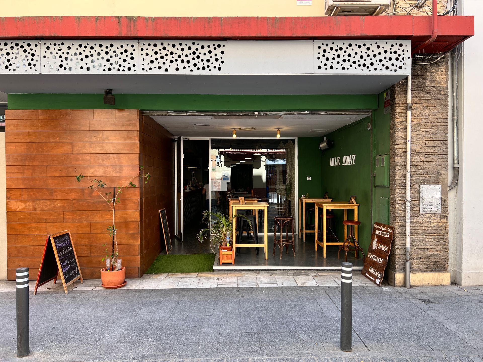 Exterior of a small restaurant with wooden accents and green trim, featuring tables inside and an outdoor menu.