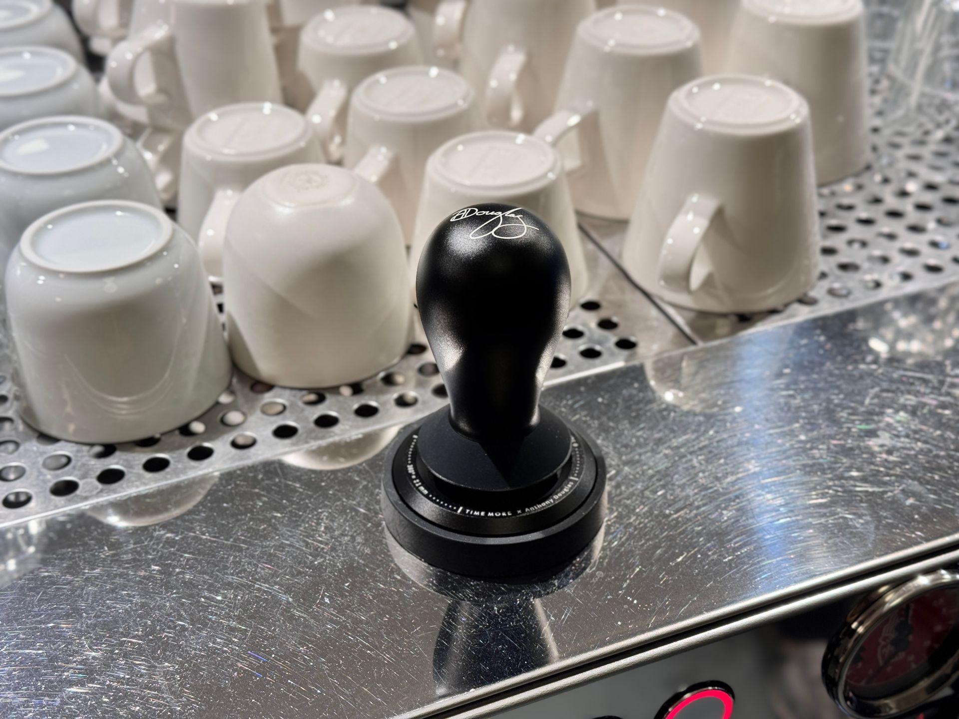 A black coffee tamper sits on the stainless steel warming tray of an espresso machine, surrounded by white cups.
