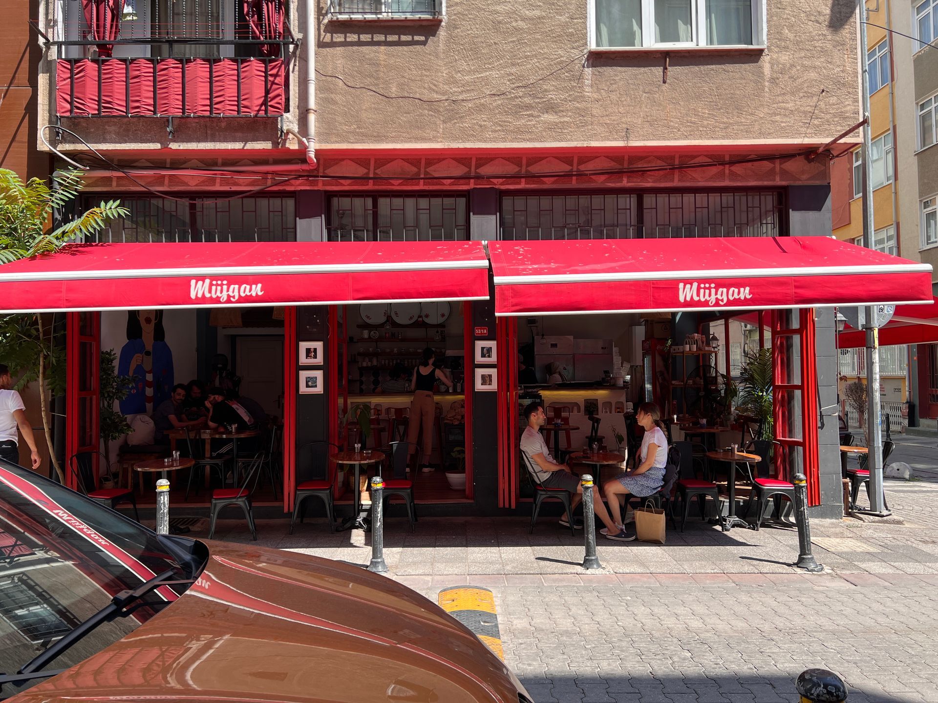 Restaurant exterior with two men seated, motorbike parked. Pink awning, white building.
