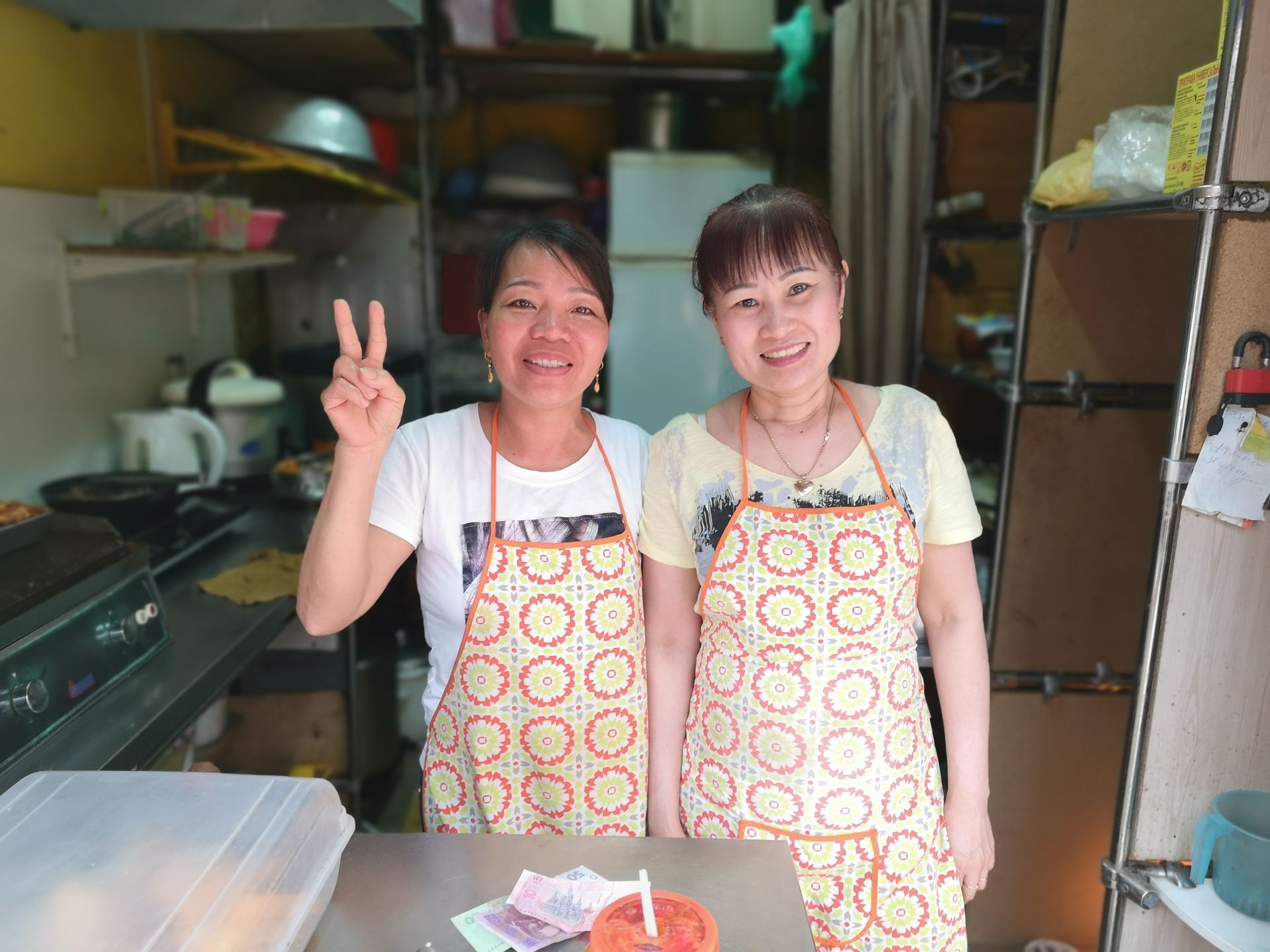 Two women in aprons smile, one giving a peace sign, standing at a small food stall, interior.