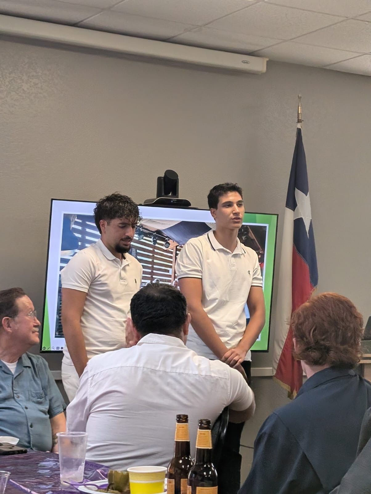 Two people in white shirts stand at the front of a meeting room beside a Texas flag and presentation screen.