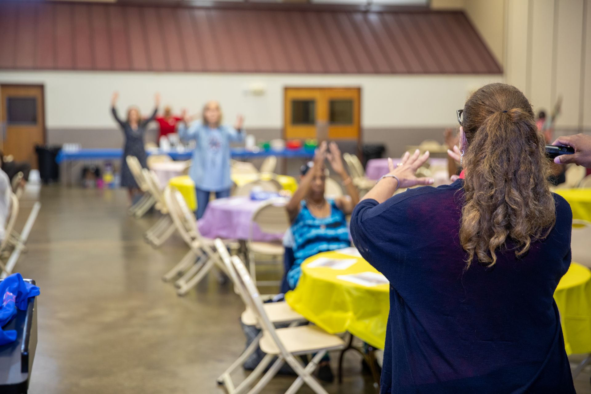 People raising hands in a bright community hall with colorful tablecloths and folding chairs