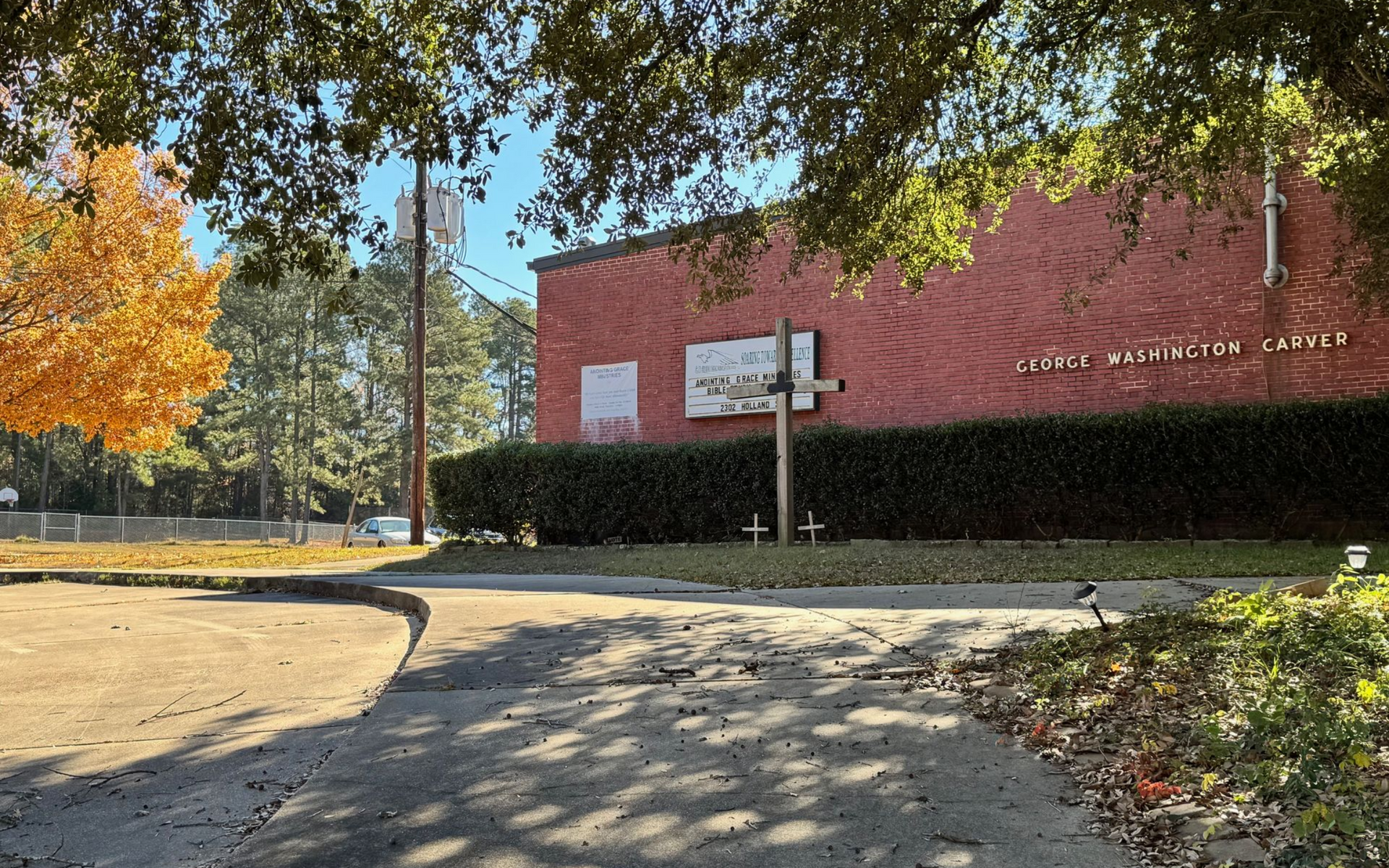 Brick building with sign and cross; trees and bushes in front on a sunny day.