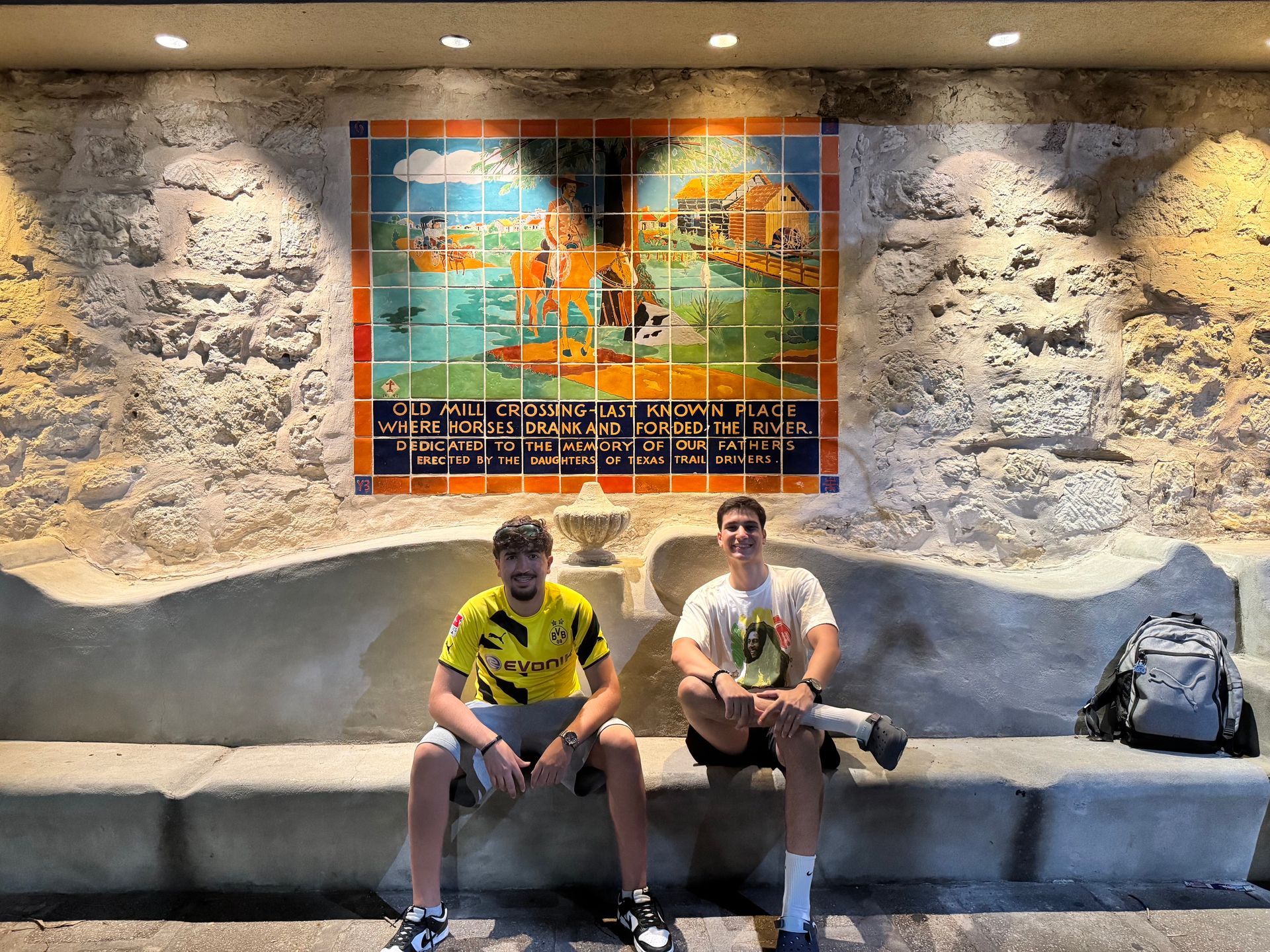 Two people sitting on a bench beneath a colorful wall mural in a lit indoor space