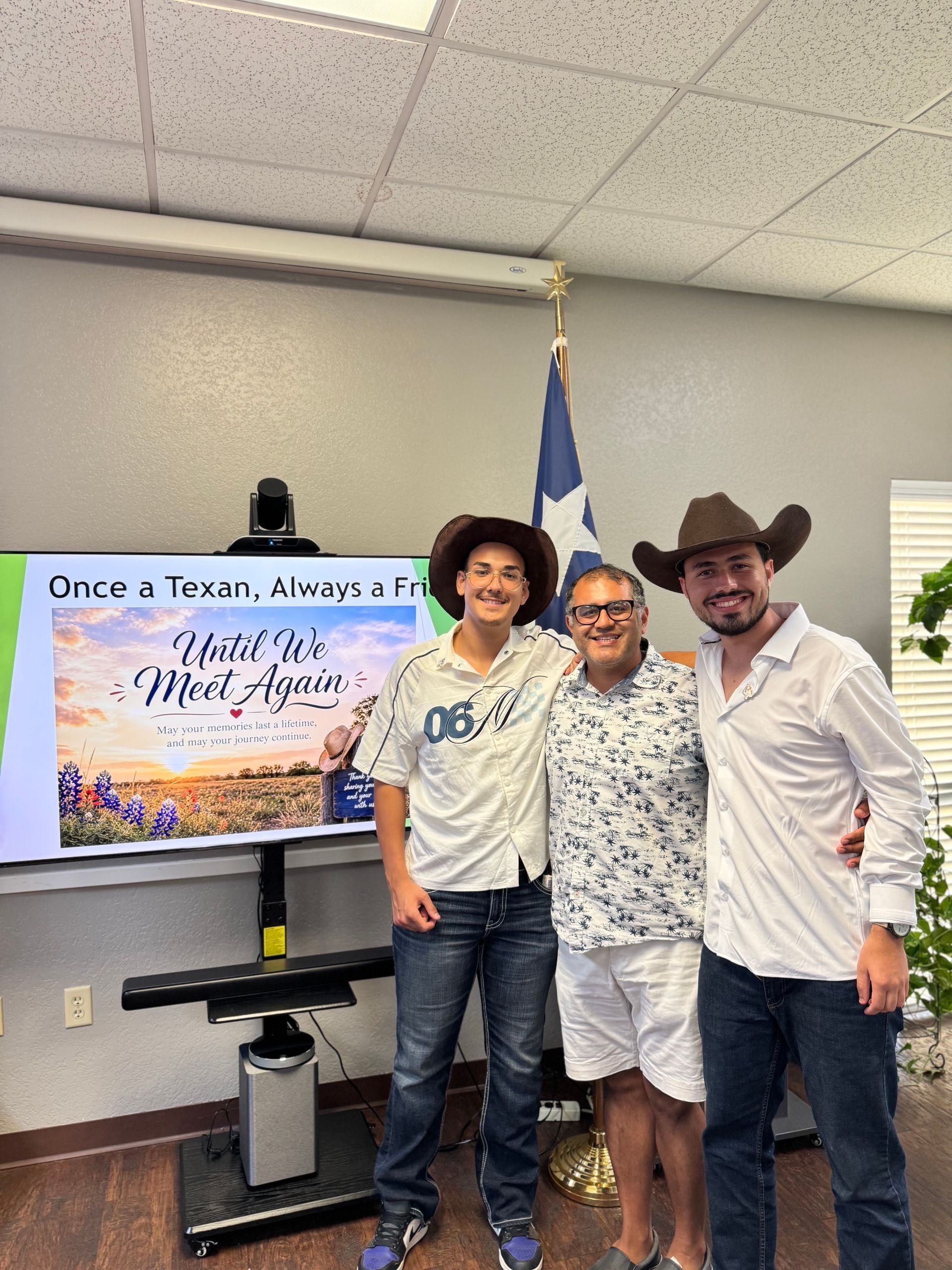 Three people posing indoors beside a screen, smiling and wearing cowboy hats and boots.