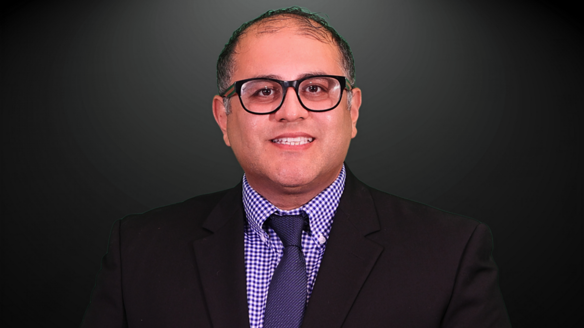 Man in a suit and glasses smiles at the camera against a dark background.