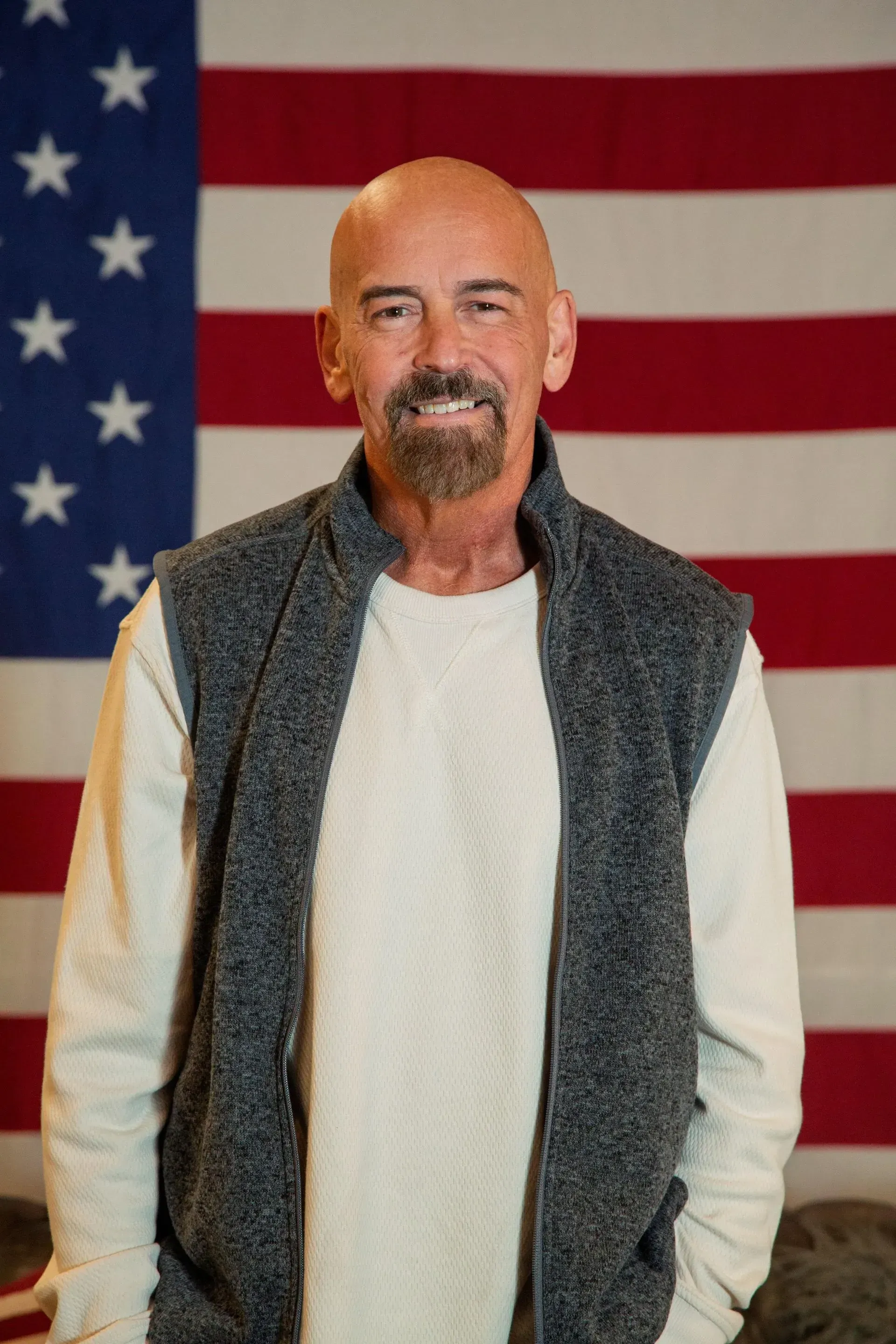 Bald man with a beard, in a vest, smiles in front of an American flag.