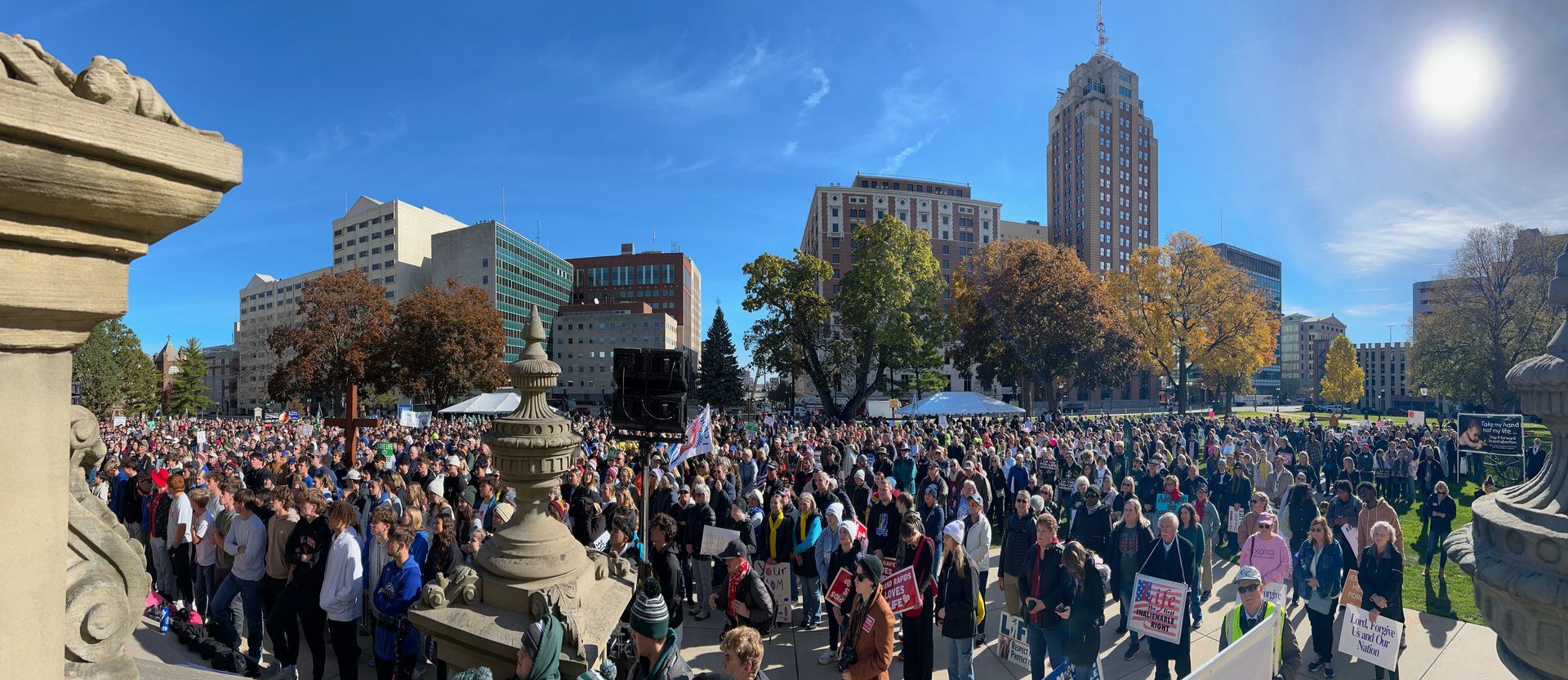 Large crowd gathered in a sunny city plaza with buildings, trees, and a tall tower in the background.