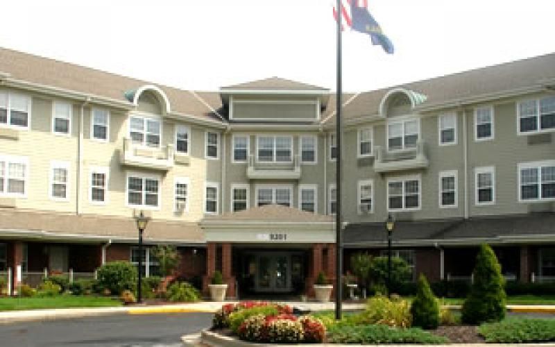 A beige, multi-story assisted living facility with a central entrance, landscaped gardens, and an American flag.