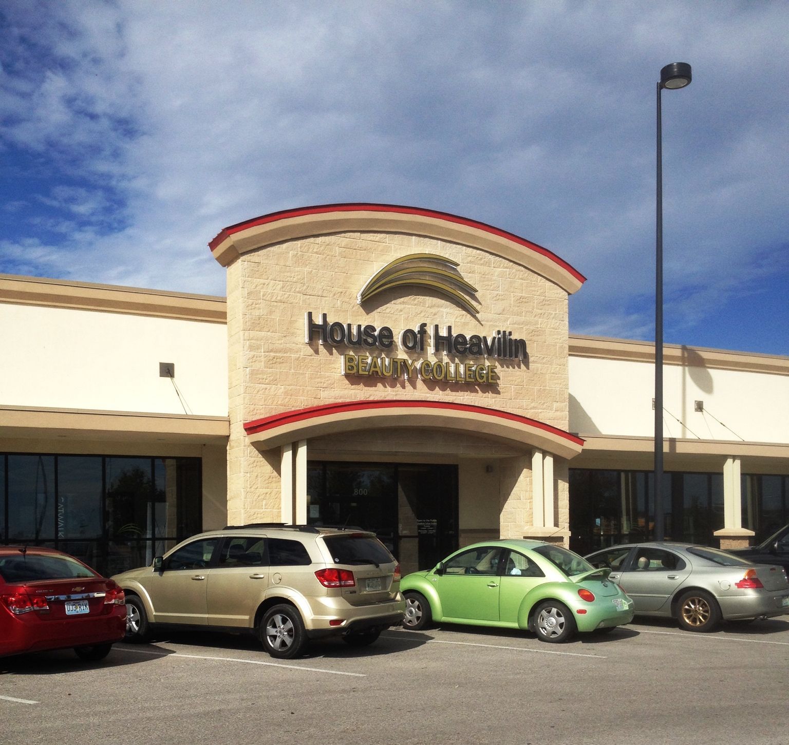 A beige storefront for House of Heavilin Beauty College with a tan arched entrance and several parked cars in the lot.
