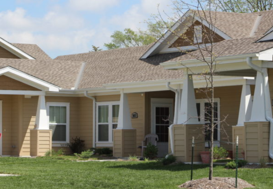 A single-story building with tan siding, a brown shingled roof, and white-trimmed porches surrounded by a grass lawn.