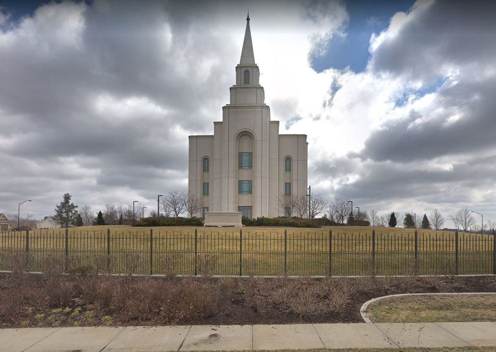 A modern white temple with a central spire, set on a raised grassy hill behind a metal fence under a cloudy sky.