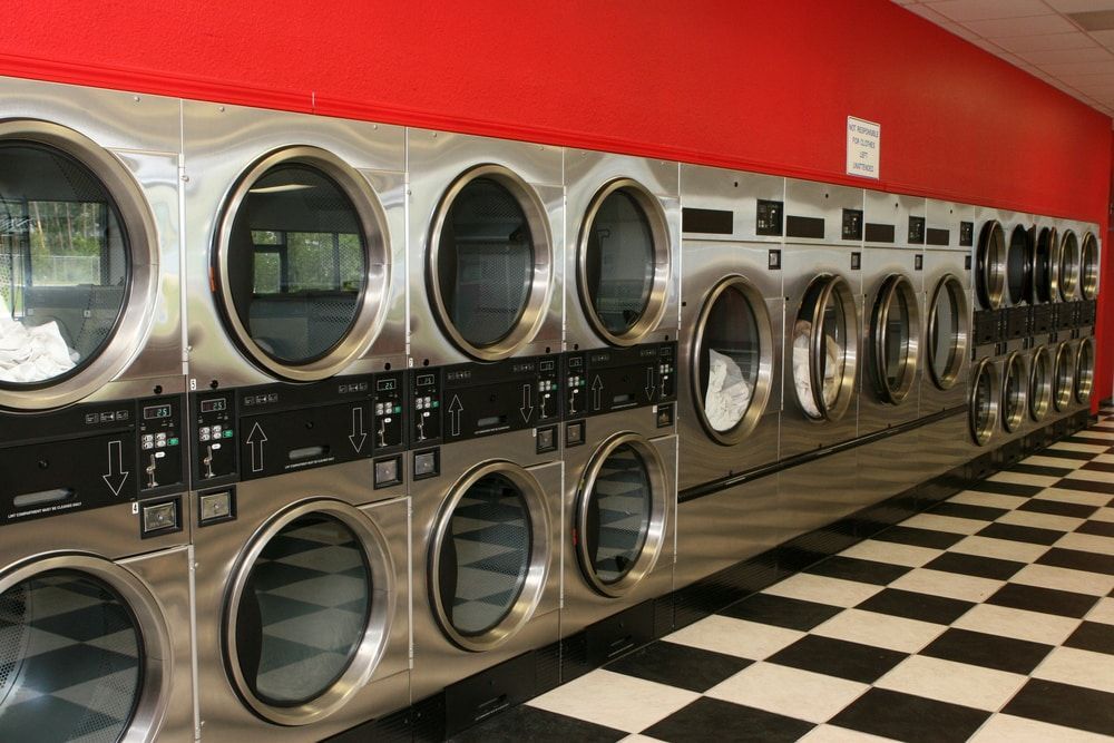 Row of stainless steel front-load washers in a laundromat with red walls and black-and-white checkered floor