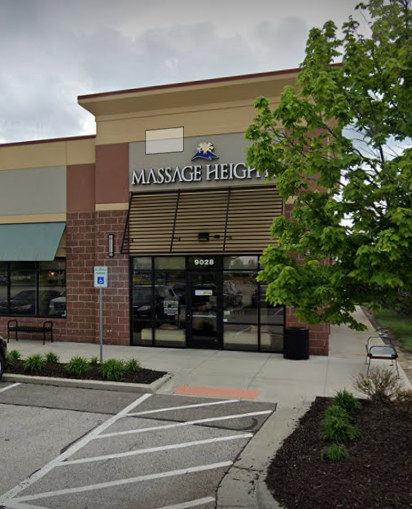 Massage Heights storefront in a tan and brick-colored shopping center with a glass entrance and a nearby tree.
