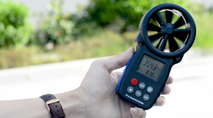 Hand holding a digital anemometer outdoors, showing its display and fan sensor.