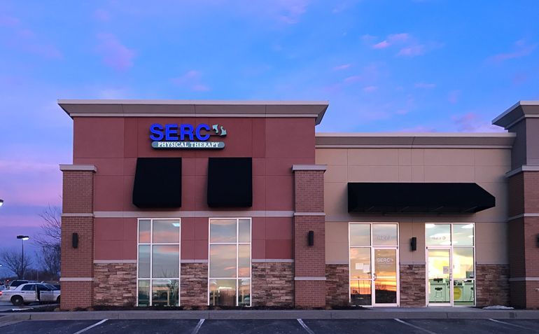 A SERG Physical Therapy building with red-brick and tan facade under a sunset sky, with black awnings above the windows.