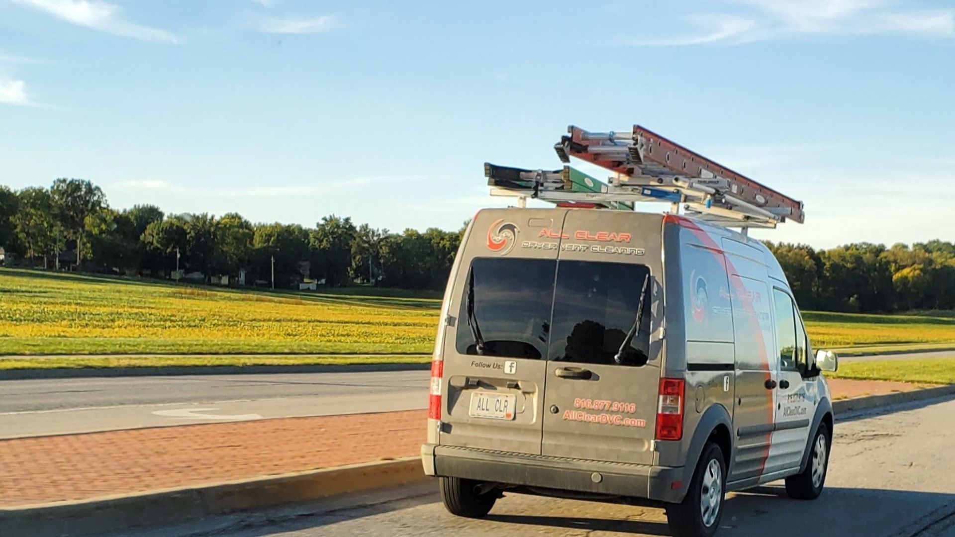 Silver work van with ladders on top driving along a road beside a grassy field at sunset