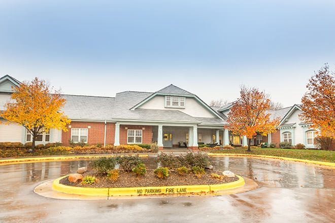 A single-story brick building with a gray roof and autumn trees, viewed across a wet circular driveway with landscaping.