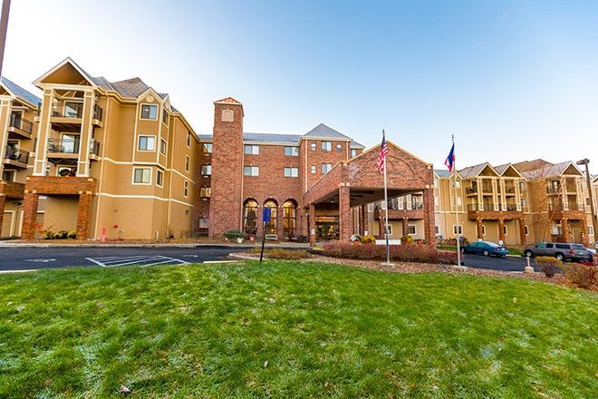 A three-story hotel or senior living facility with tan stucco and brick siding, a porte-cochère, and a green front lawn.