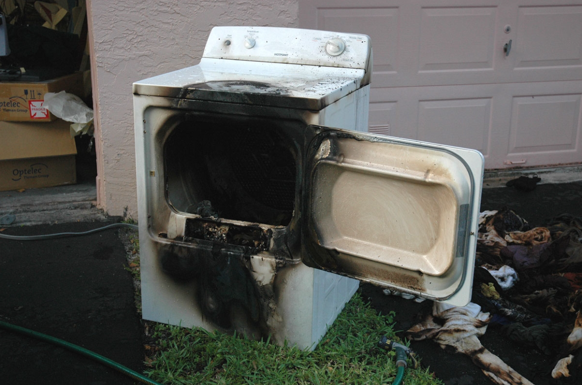 Burned white washing machine with open door, charred inside, sitting outdoors by a garage.