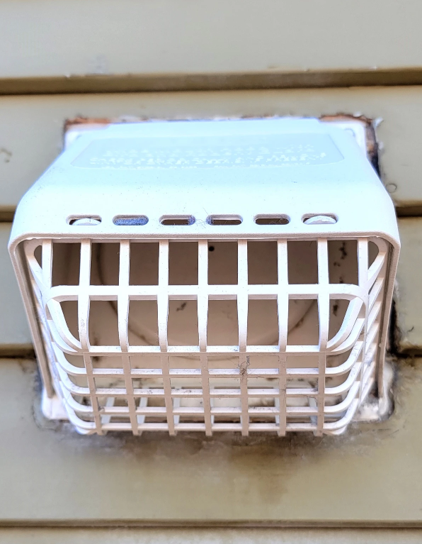 White wall-mounted vent or speaker grille on beige siding, with a protective cage over the front