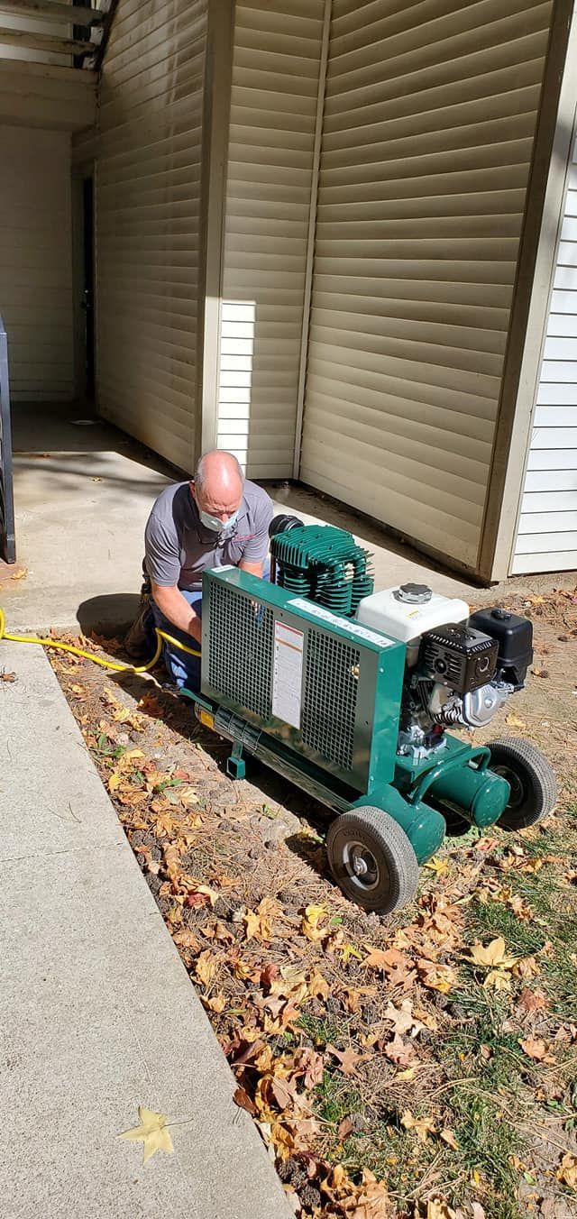 Man kneeling beside a green utility cart, clearing leaves along a sidewalk by a building.