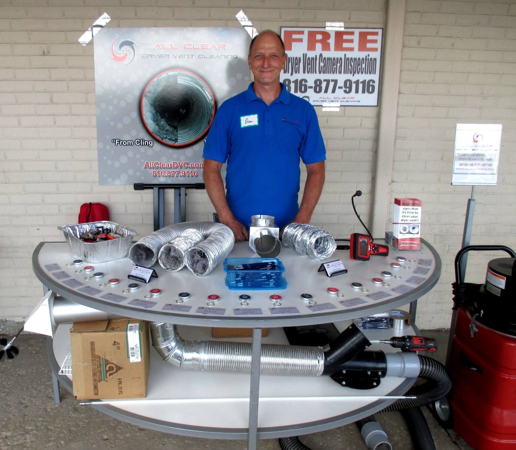 Man in blue shirt standing behind display table of shiny metal duct parts and tools at an outdoor booth