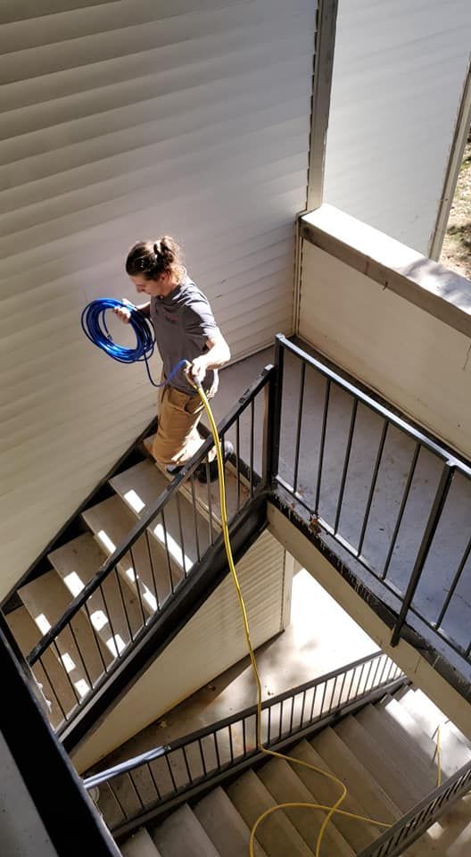 Person standing on a stair landing holding a blue hose near a sunlit stairwell
