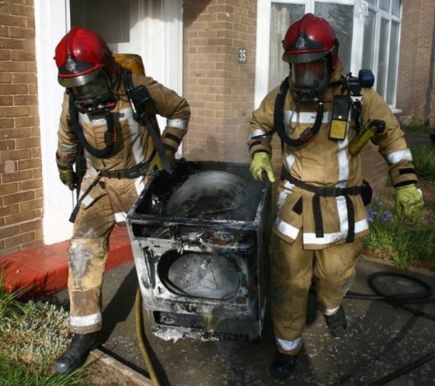 Two firefighters in tan gear stand beside a burned appliance outside a brick building.