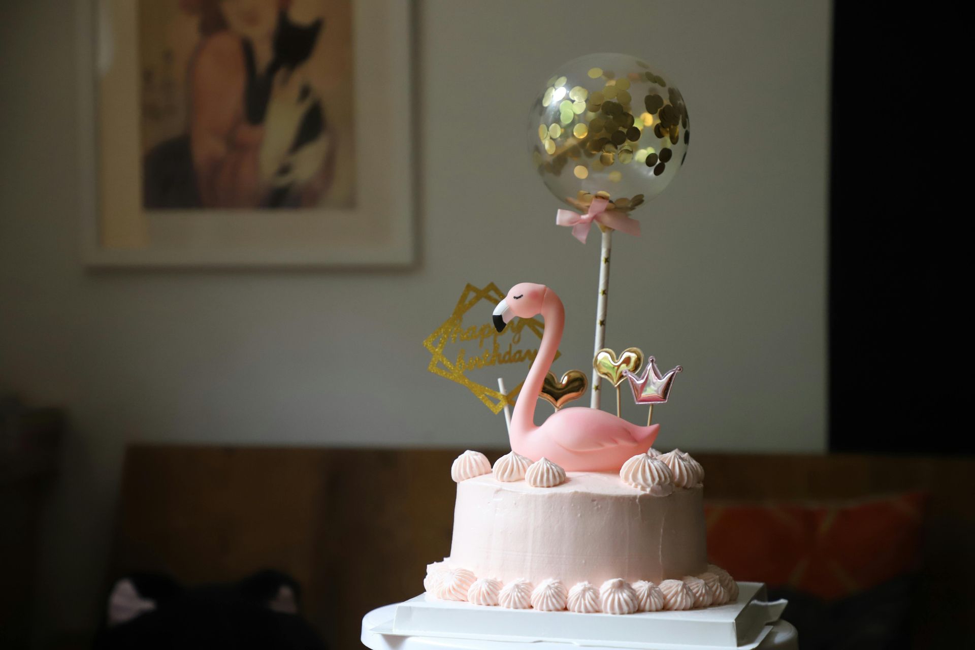 A pink cake topped with a flamingo figurine, a small confetti balloon, and decorative gold charms.